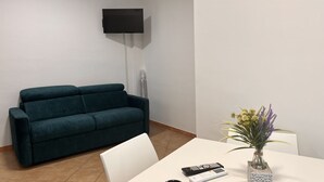 Ein Zimmer mit einem türkisfarbenen Sofa, einem weißen Tisch mit Blumen und einem an der Wand befestigten Fernseher.
