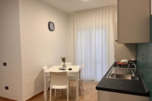 A compact kitchen with a dining table, chairs, a clock, and a window with curtains.