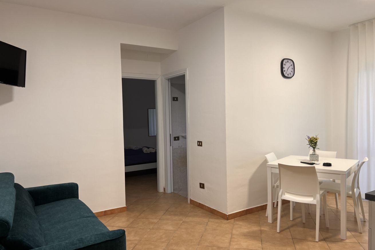 Een moderne woonkamer met een blauwgroene bank, een eethoek met witte stoelen, een kleine tafel en een klok aan de muur.