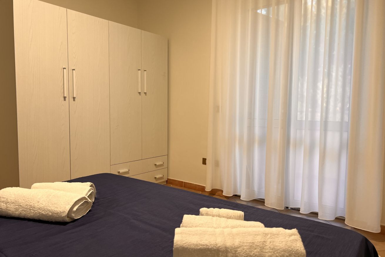 Un dormitorio con cama, dos toallas blancas, armario y ventana con cortinas traslúcidas.