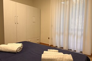 Un dormitorio con cama, dos toallas blancas, armario y ventana con cortinas traslúcidas.