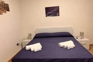 Ein ordentlich bezogenes Bett mit zwei zusammengerollten Handtüchern, ein Nachttisch mit einer Lampe und ein gerahmtes Bild einer Brücke an der Wand.