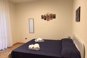 Een hotelkamer met een bed, twee opgerolde handdoeken, een kunstwerk aan de muur en een raam met gordijnen.
