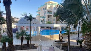 A pool area with a clear blue pool, palm trees, and a multi-story building in the background. - Mamad Panoramic Red Sea | Chic 3BR Haven (Eilat)