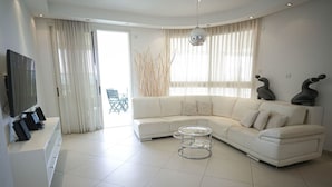 Un salon moderne avec un canapé d’angle blanc, une table basse en verre et un téléviseur à écran plat posé sur une console blanche.