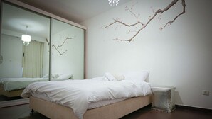 A bedroom with a bed, a small bedside table, a mirror, and a wall-mounted branch decoration. - Mamad Panoramic Red Sea | Chic 3BR Haven (Eilat)