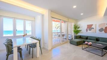 A modern living room with a glass table, green chairs, a green sofa, and a view of the ocean through large windows.