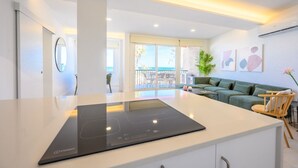 Une cuisine moderne dotée d’une plaque de cuisson à induction intégrée, d’une grande fenêtre offrant une vue sur l’extérieur et d’un coin salon confortable avec un canapé vert et des chaises en bois.