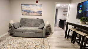 A living room with a grey sofa, two side tables with lamps, a patterned rug, a painting of birds and water, a kitchen area with a TV displaying Netflix, and a small potted plant. - Ocean Oasis - Beachfront Condo with breathtaking views (Hampton)