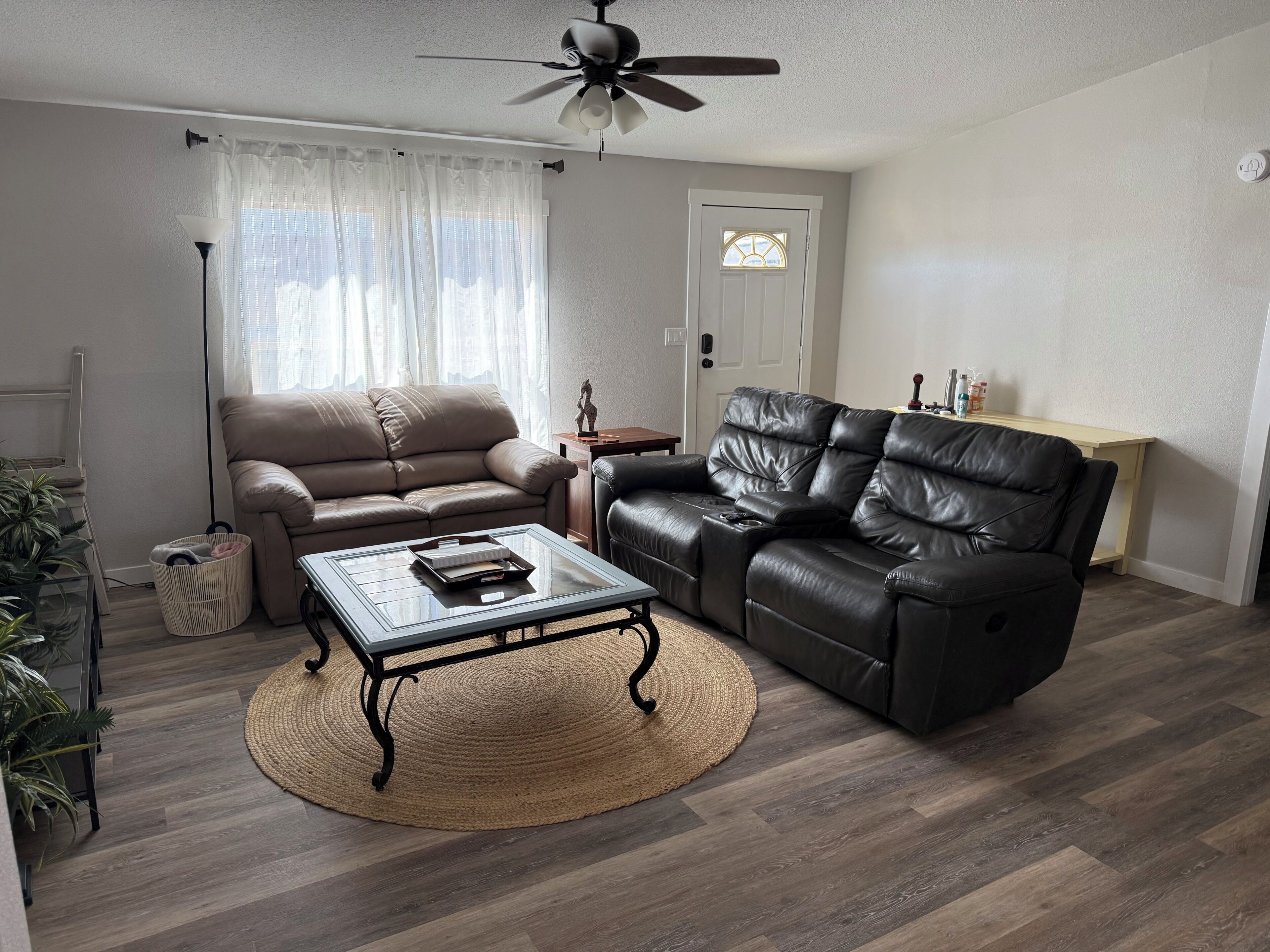 A living room with a sofa, a recliner, a coffee table, a ceiling fan, a door with a window, and a small table with decorative items.