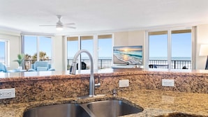A kitchen with granite countertops, a stainless steel sink, a ceiling fan, and a TV displaying a sunset view.