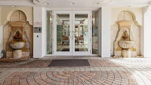A hotel entrance with a brick-paved driveway, double doors, and two stone fountains on either side.
