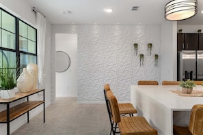 Una cocina moderna con pared blanca texturizada, un refrigerador integrado y una zona de comedor con sillas de madera y una mesa blanca.