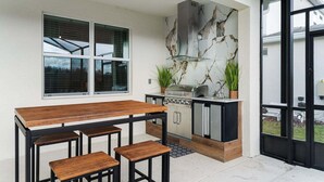 Eine Außenküche mit Holztisch und -stühlen, einem Grill und einer Rückwand mit Marmoroptik.