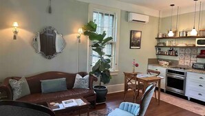Una sala de estar con un sofá de cuero marrón, una mesa de centro y un comedor con una mesa y sillas de madera. Hay una planta grande en maceta, un espejo en la pared y una cocina con microondas y horno.