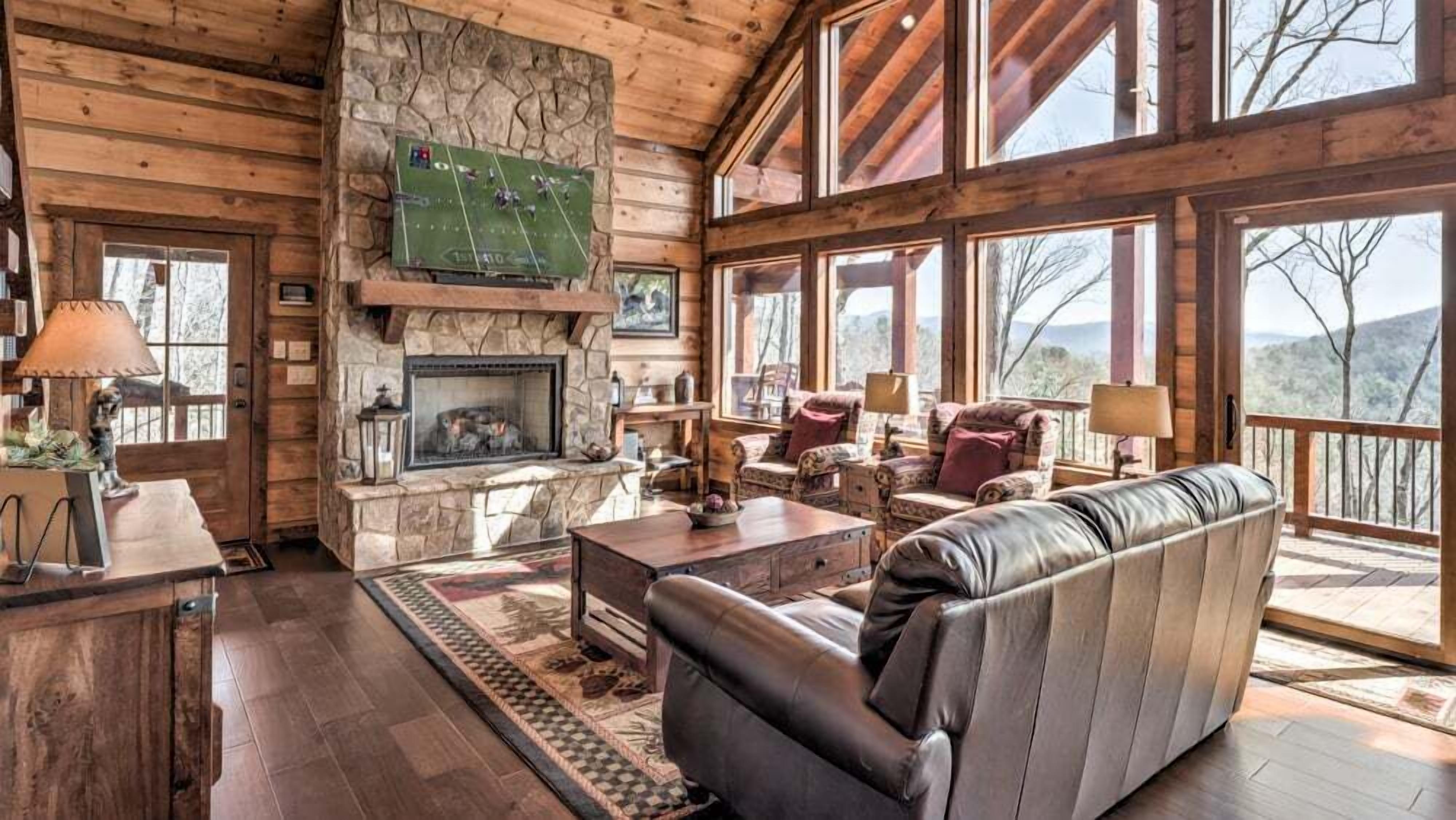 A cozy cabin interior with a stone fireplace, leather sofa, and large windows offering a view of the outdoors.