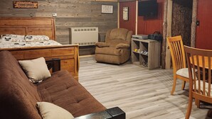 A cozy room with a wooden ceiling, a bed with a patterned duvet, a brown sofa, a wooden dining table with chairs, and a small TV on the wall.