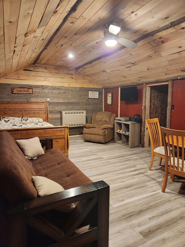A cozy room with a wooden ceiling, a bed with a patterned duvet, a brown sofa, a wooden dining table with chairs, and a small TV on the wall.