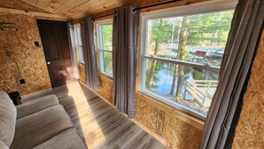 A cabin interior with a wooden ceiling, a sofa, and large windows overlooking a natural landscape.