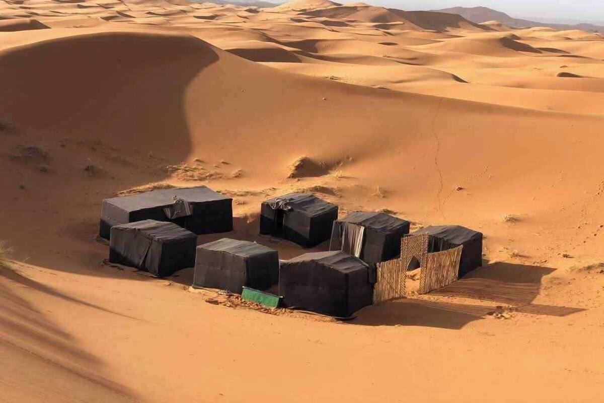 A desert landscape with sand dunes and a group of black structures, possibly tents or temporary shelters, arranged in a semi-circle.