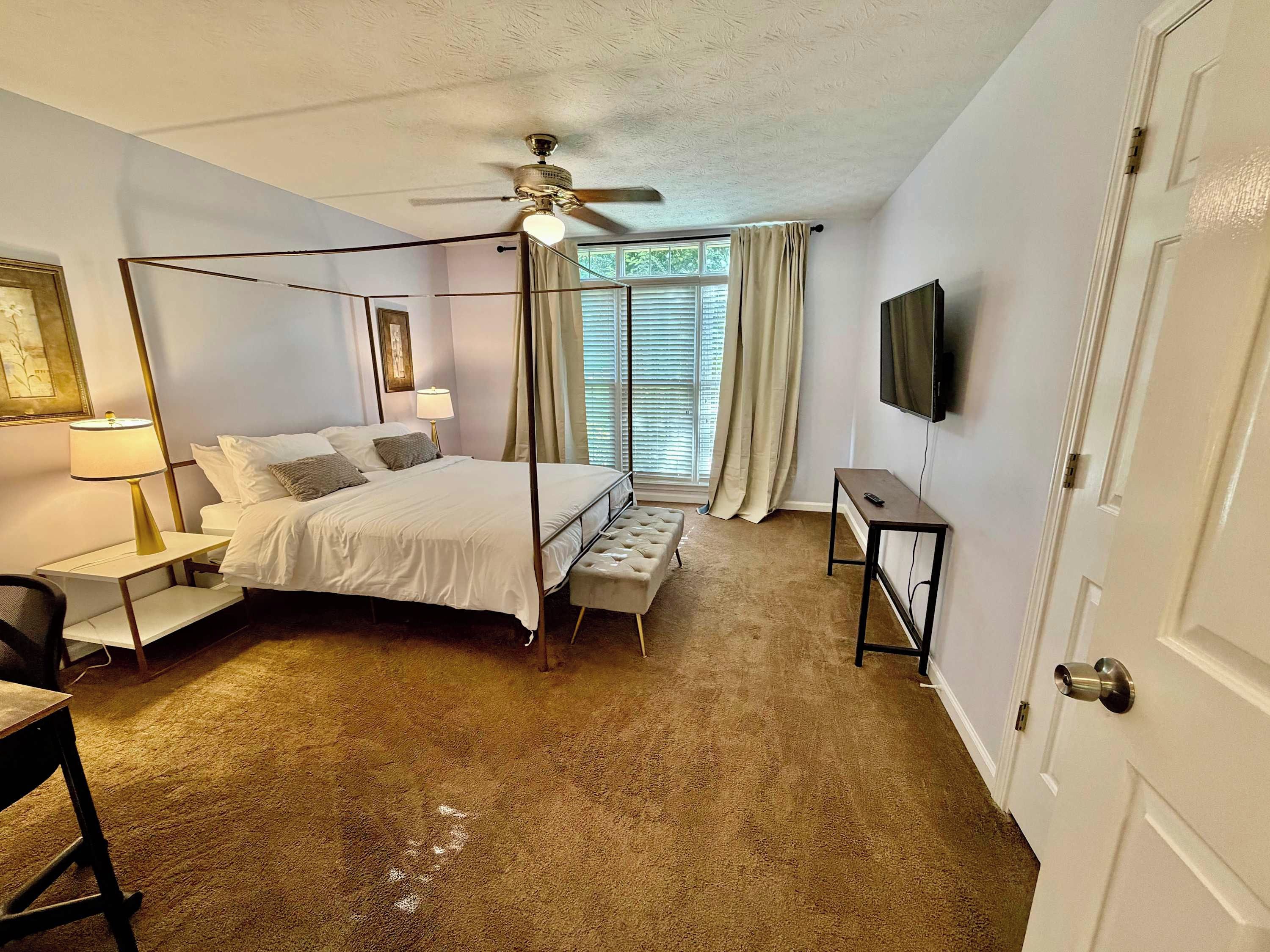 A hotel room with a four-poster bed, a desk, a chair, a ceiling fan, a television, and a window with blinds.
