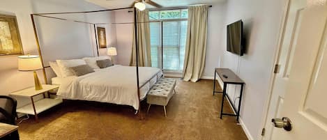 A hotel room with a four-poster bed, a desk, a chair, a ceiling fan, a television, and a window with blinds.