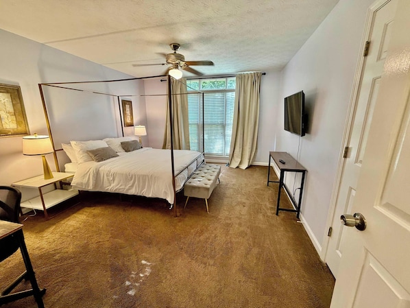 A hotel room with a four-poster bed, a desk, a chair, a ceiling fan, a television, and a window with blinds.