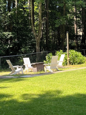 Une zone herbeuse avec trois chaises Adirondack blanches et une table de pique-nique, le tout entouré d’une clôture en treillis métallique et d’arbres denses.