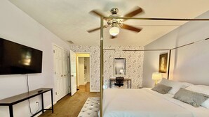 Una habitación de hotel con cama, escritorio, televisor, ventilador de techo y baño visible a través de una puerta abierta.