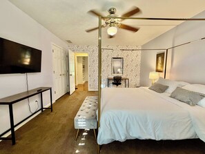 Una camera d'albergo con un letto, una scrivania, un televisore, una ventola a soffitto e un bagno visibile tramite una porta aperta.
