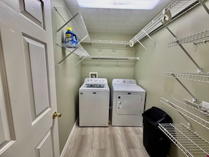 Une buanderie équipée de deux machines à laver, d’un sèche-linge, d’étagères en métal et d’une poubelle.