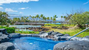 A pool with a rocky edge, surrounded by greenery and palm trees, with a view of a beach and ocean in the background. - Kolea Villas 15M, Luxury, Direct Beach Access (Waikoloa)