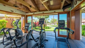 A covered outdoor gym with treadmills, elliptical machines, and a stationary bike, overlooking a landscaped garden and a clear view of the surrounding area. - Kolea Villas 15M, Luxury, Direct Beach Access (Waikoloa)