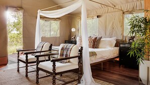 A four-poster canopy bed with a view of greenery outside, a wooden chair, a bedside table with a lamp, and a potted plant.