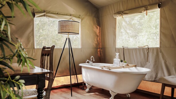 A room with a white clawfoot bathtub, a wooden chair, a table, and a floor lamp.