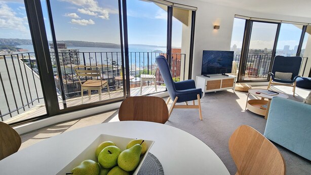 A modern living room with a large balcony, a television, and a dining area with a fruit bowl.