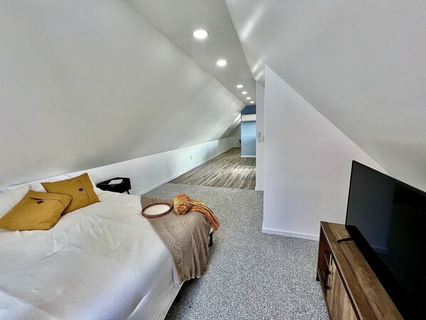 A modern, compact bedroom with a sloped ceiling, a bed with white bedding and two pillows, a small bedside table, a TV stand, and a wooden floor.