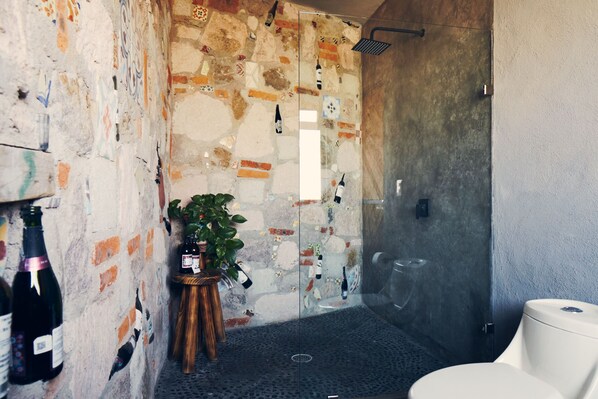 Un baño con pared de piedra, ducha, inodoro y una pequeña mesa con una planta y botellas de vino.