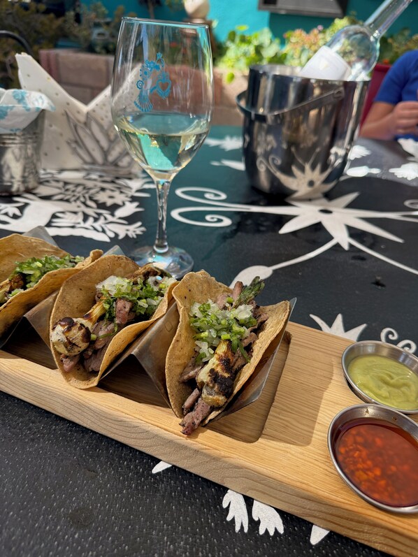Tres tacos sobre una tabla de madera, una copa de vino blanco y dos recipientes pequeños de salsa.