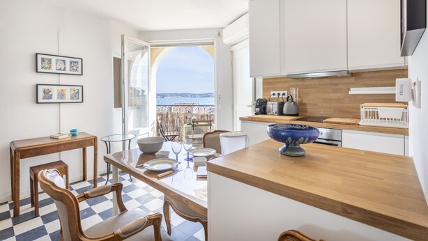 Une cuisine moderne avec des meubles blancs, un plan de travail en bois et une vue sur la ville par une porte ouverte.