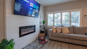 Una sala de estar con una televisión de pantalla plana montada en la pared, una chimenea debajo, un sofá con cojines y una ventana con persianas.