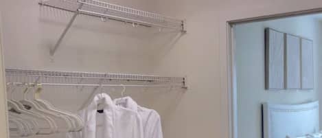 A room with a white robe hanging on a rack, a bed with white bedding, and a small bedside table with a lamp.