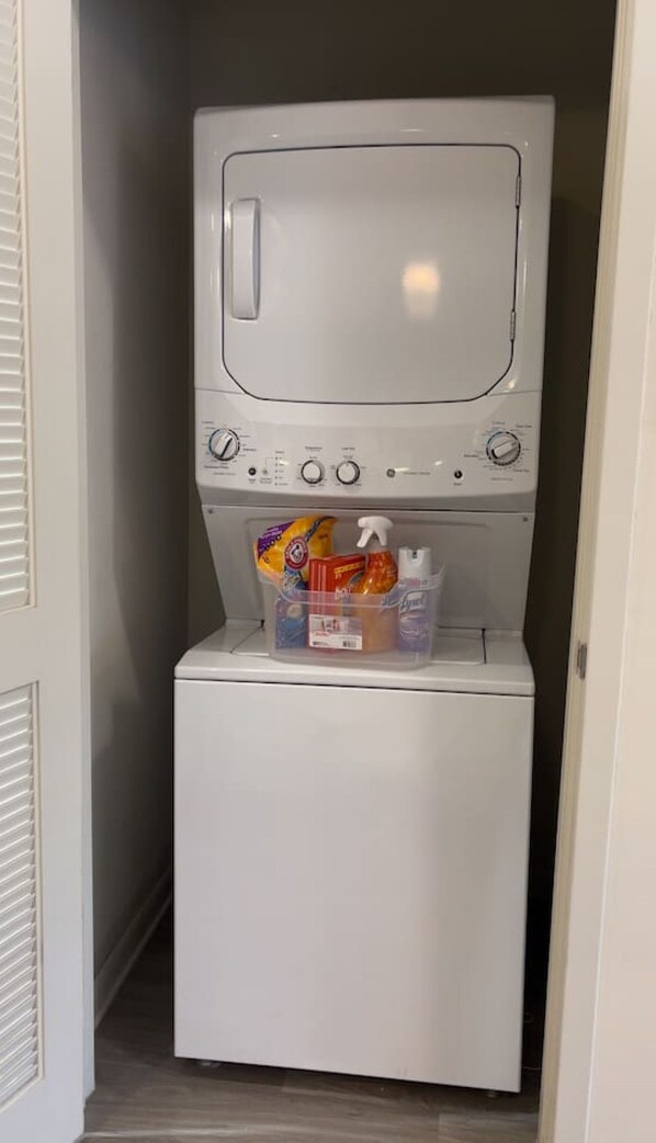 Une machine à laver compacte, de couleur blanche, avec une porte à chargement frontal, un panneau de commande doté de plusieurs boutons et un compartiment de rangement situé en dessous, contenant des produits de nettoyage.