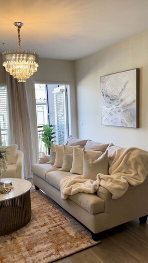 A living room with a beige sofa, a round coffee table, a chandelier, and a large window with blinds. - Atlanta Beltline Luxury Suite (Atlanta)