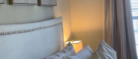 A bedroom with a bed, pillows, a headboard, a nightstand, a lamp, and a window with curtains.