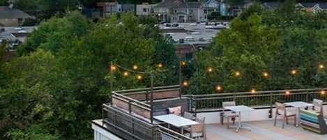 A rooftop terrace with outdoor seating and string lights, overlooking a cityscape at dusk.