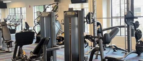 Un gimnasio con diversas máquinas de ejercicio, una alfombra estampada y ventanas amplias.