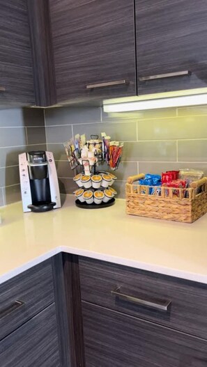 A kitchen with dark wooden cabinets, a coffee maker, a utensil holder, and a basket of snacks.