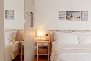 Et soveværelse med to senge, et natbord med lampe, en vægmonteret hylde og tre billeder i rammer.