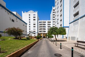 Ein Weg führt zu einem mehrstöckigen Gebäude, einer grünen Fläche mit Bäumen und einem wolkenlosen, blauen Himmel.